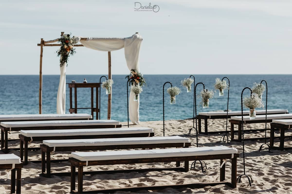 Pedregal beach cabo wedding ceremony: wooden arch with white drape, benches on the sand, ocean horizon