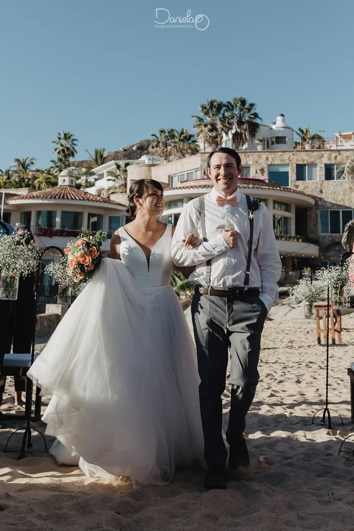 Bride and groom walking the sandy aisle after a Pedregal beach cabo wedding, Villa Marcella and shepherd-hook florals behind