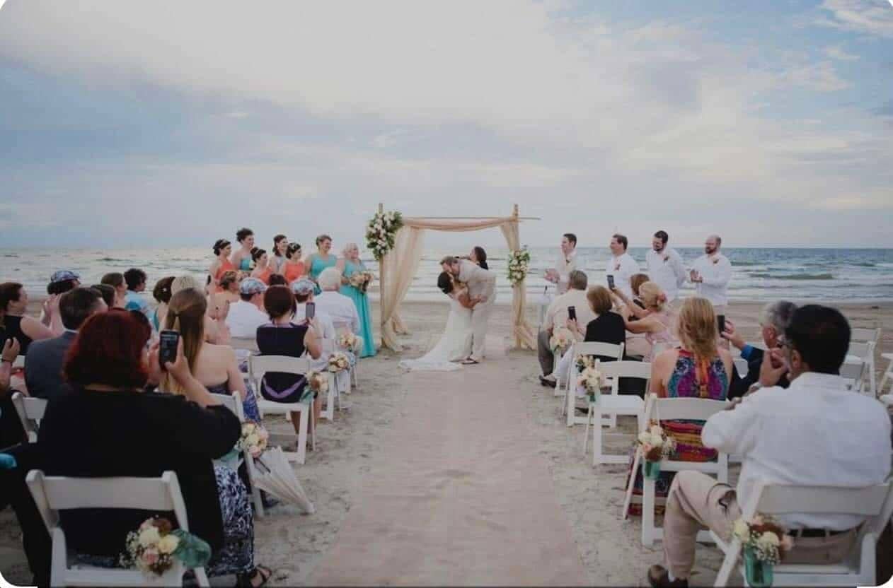 First kiss under a floral arch at a Casa Mareva cabo wedding ceremony on the beach, guests filming from white chairs